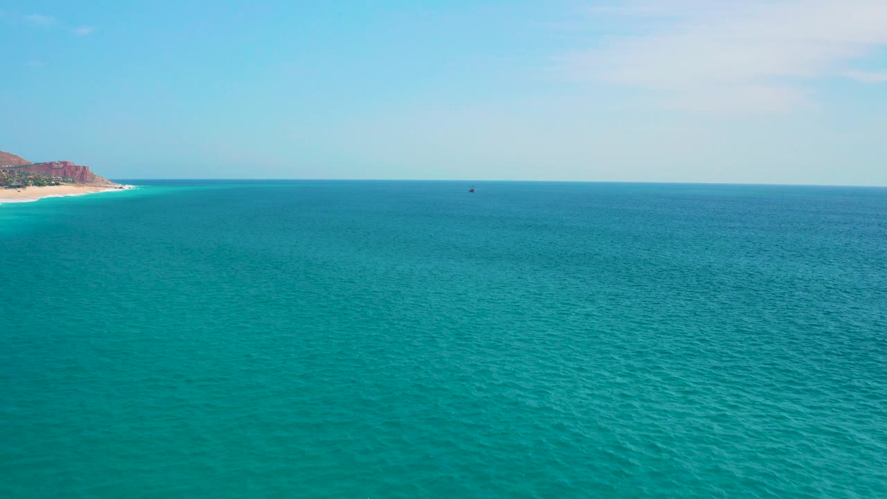 The ocean remains dominant, with a small dark offshore object near the horizon, the left shoreline barely entering frame, and no visible people.
