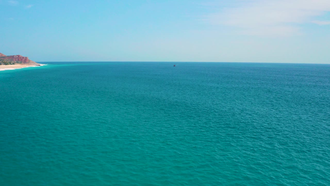 Wide aerial view at Secrets Marquis Los Cabos, Mexico, showing turquoise ocean, pale sky, a small left-edge shoreline and rocky hill, with no visible people.