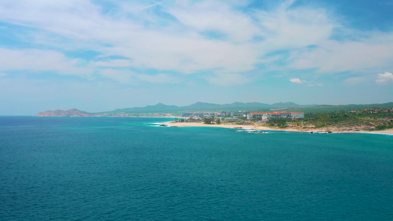 Wide aerial frame over turquoise water toward the Secrets Marquis Los Cabos shoreline in Mexico, with surf, resort buildings, distant mountains, cloudy sky, and no visible people.