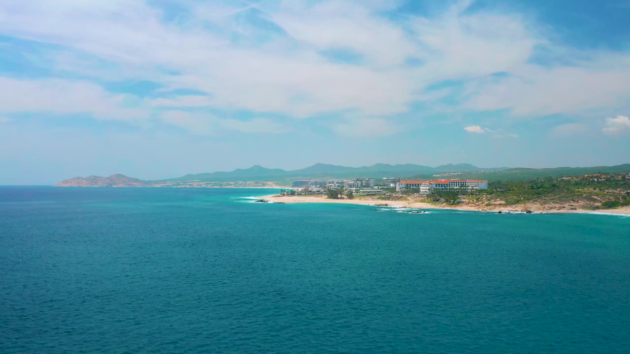 Elevated view across the ocean toward a pale beach at Secrets Marquis Los Cabos, with low buildings near the shore, mountains on the horizon, and no visible people.