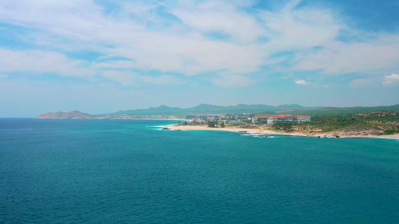 A wide aerial coastal frame shows blue-green water in front, a bright surf line at the beach, resort buildings to the right, distant landforms to the left, and no visible people.
