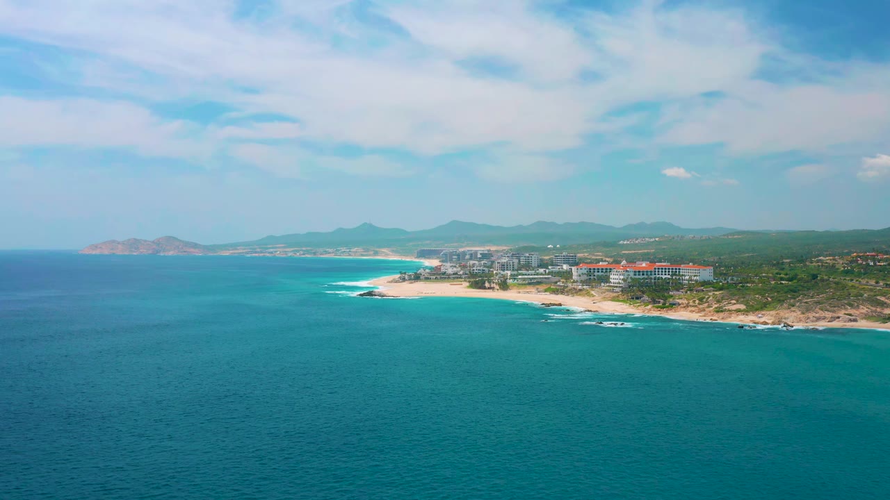 At 0:01, the aerial frame holds open turquoise water in the foreground, white surf along the beach, resort buildings on the coast at Secrets Marquis Los Cabos, Mexico, and no visible people.