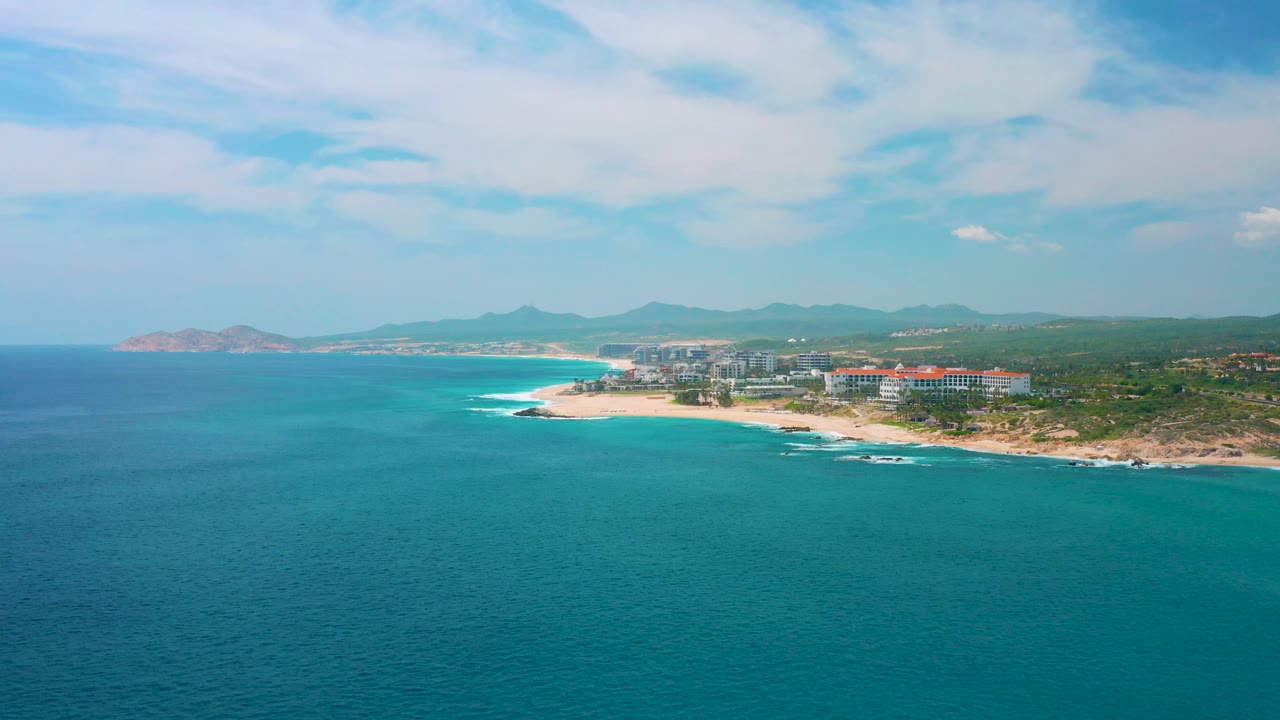 At 0:02, the coastline curves through the frame with turquoise sea, a bright sandy beach, resort buildings at Secrets Marquis Los Cabos, Mexico, mountain ridges, and no visible people.