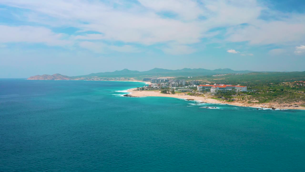 At 0:03, blue-green ocean leads to a pale beach and low surf, with the resort area and coastal hills of Secrets Marquis Los Cabos, Mexico in the distance and no visible people.