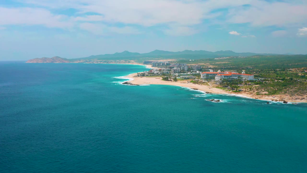 Wide aerial frame over blue-green ocean toward the Secrets Marquis Los Cabos shoreline, with pale beach, low waves, resort blocks, inland greenery, distant hills, and no visible people.