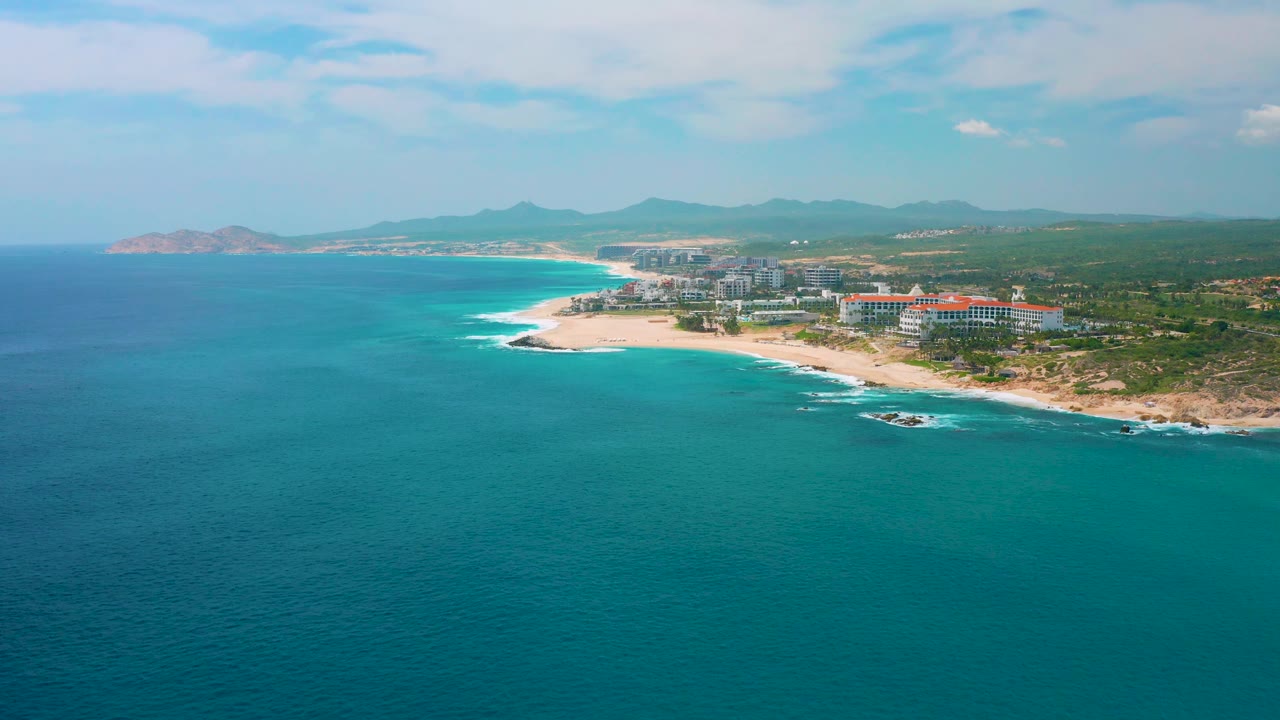 Aerial coastal view at Secrets Marquis Los Cabos, Mexico showing a broad ocean foreground, white surf on the sand, a resort complex beside the beach, a long coastline, hazy hills, and no visible people.