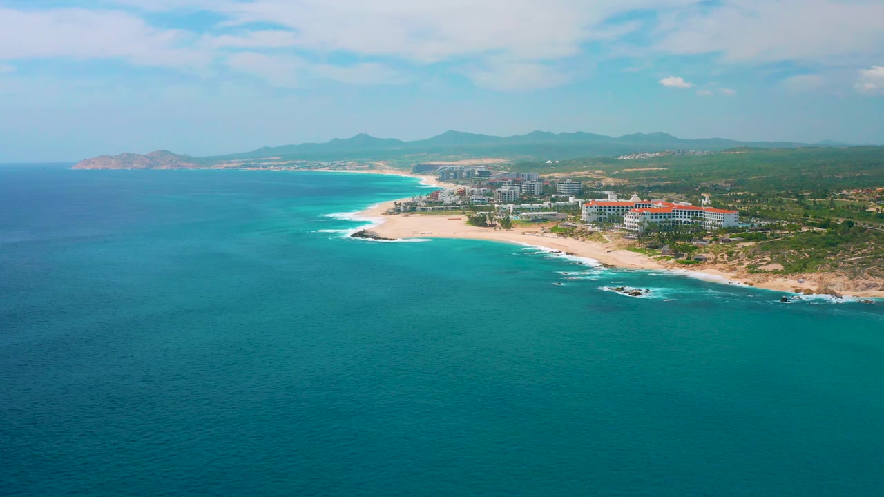 Drone-style frame looking across turquoise sea toward a sandy point and hotel buildings at Secrets Marquis Los Cabos, with inland greenery, distant ridges, scattered clouds, and no visible people.