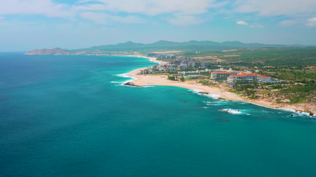 Aerial view of turquoise ocean, sandy beach, resort buildings, and distant hills.