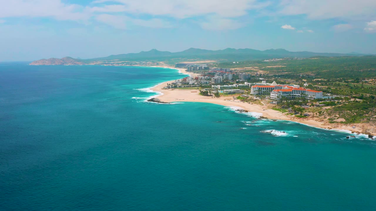 Wide aerial frame of Secrets Marquis Los Cabos with blue-green water occupying most of the foreground, surf along the beach, resort blocks to the right, and no visible people.