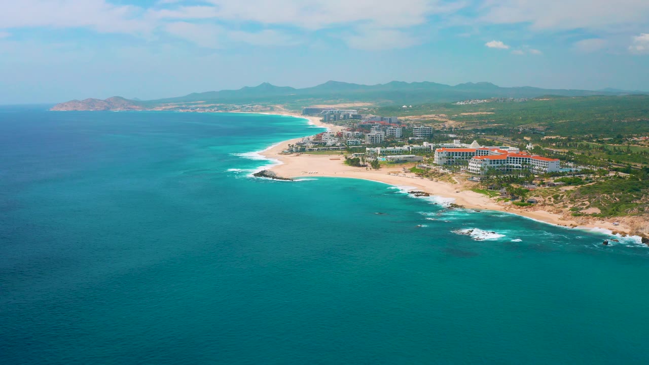 Aerial view of the Mexico resort shoreline with clear blue ocean, white wave lines over rocks near shore, resort buildings on the right, and no visible people.