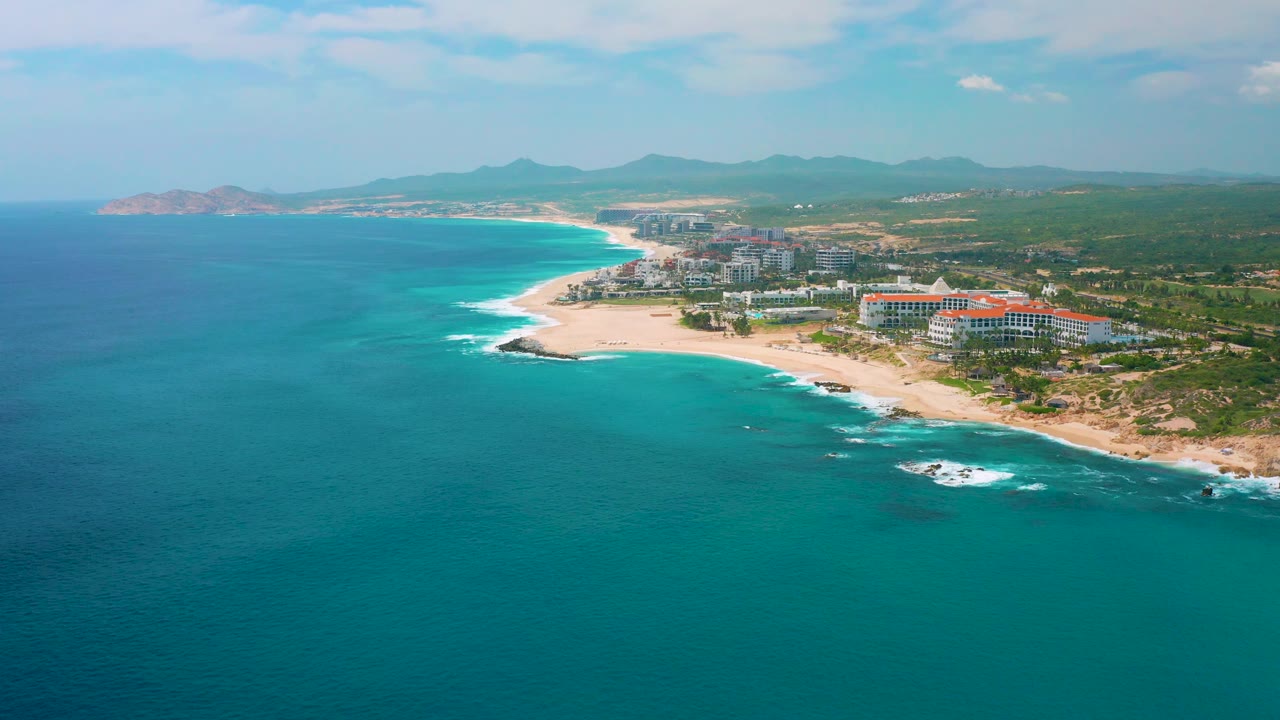 Drone-supported view of Secrets Marquis Los Cabos showing the ocean foreground, a beach curve leading toward a distant headland, resort buildings at right, and no visible people.