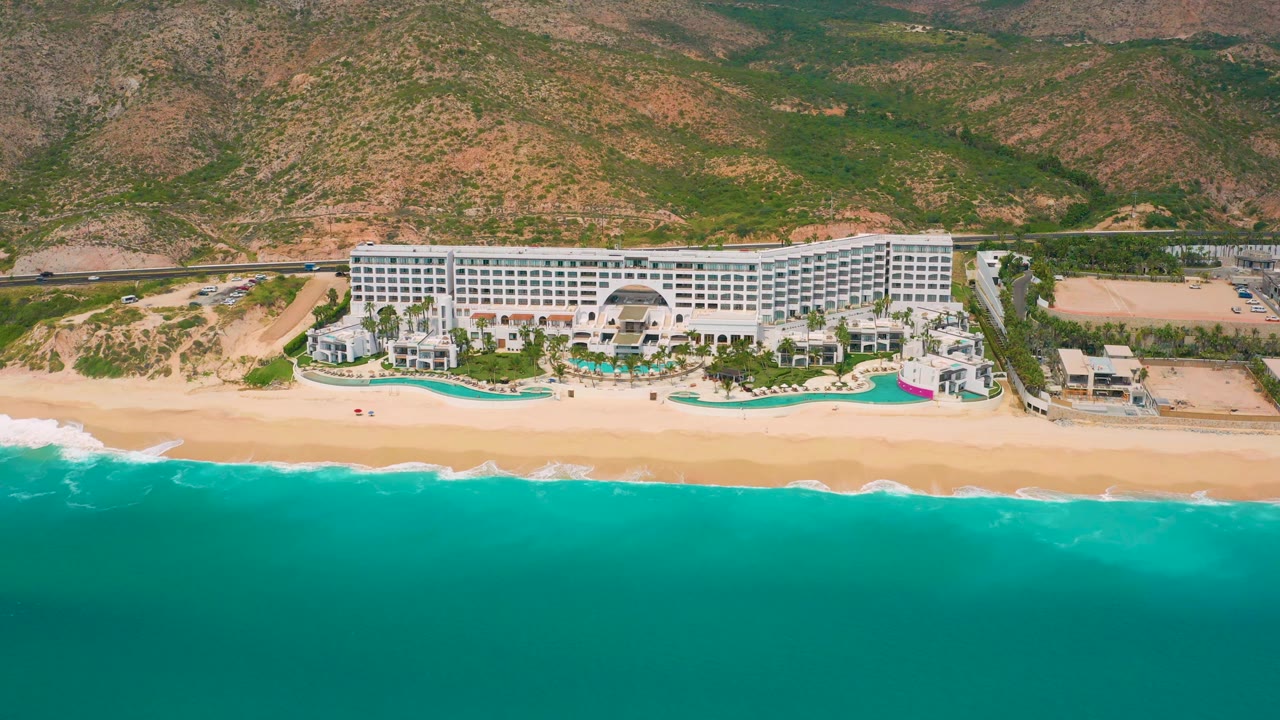 A wide aerial view of Secrets Marquis Los Cabos shows the white hotel block centered above the beach, turquoise pools along the resort edge, teal water in the foreground, and no visible people.