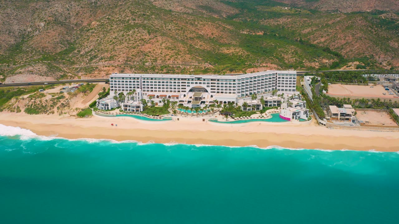 The aerial composition holds on the Mexico beachfront resort as a brighter surf band meets the sand, with pool terraces and the long white hotel backed by dry green-brown hills and no visible people.