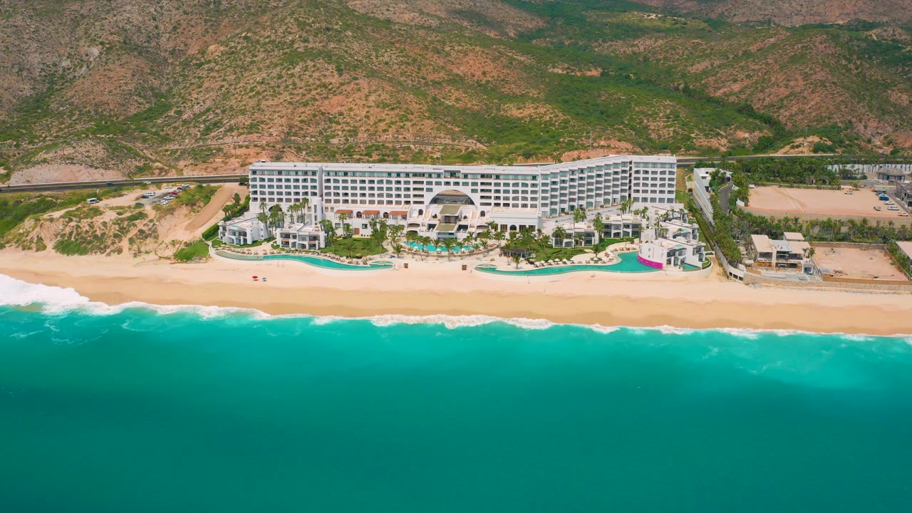 Aerial resort view with ocean foreground, sandy shore, large white hotel, and hills.