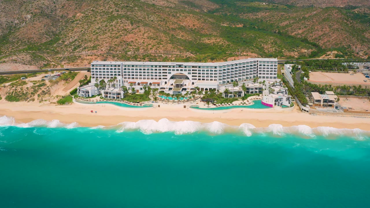 Aerial view of Secrets Marquis Los Cabos in Mexico, showing the resort centered above the beach as white surf spreads along the sand and no visible people appear in the frame.