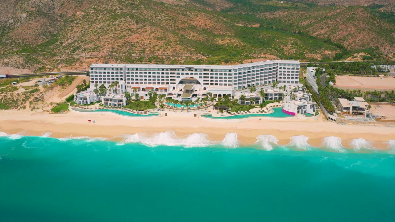 Large white resort behind a beach with waves and green-blue ocean water.