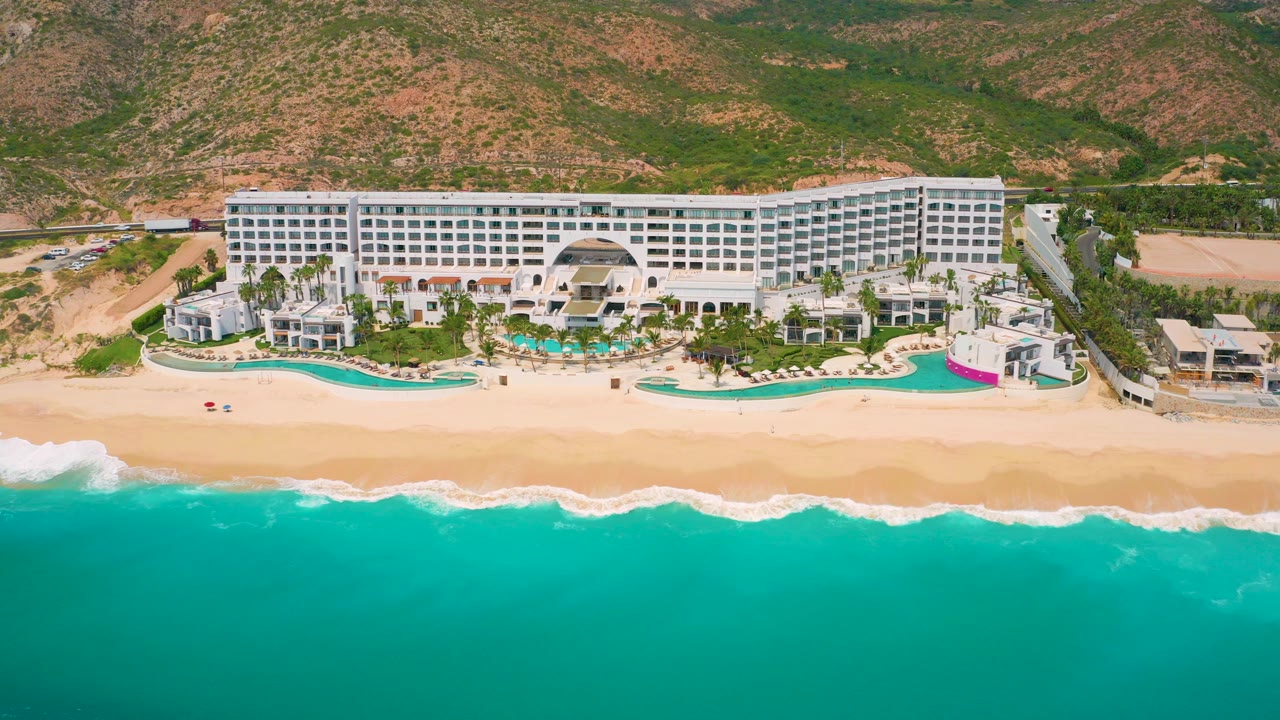Aerial frame of Secrets Marquis Los Cabos in Mexico with turquoise water in the foreground, a sandy beach, resort pools, white hotel buildings, hillside terrain, and tiny visible people near the shore.