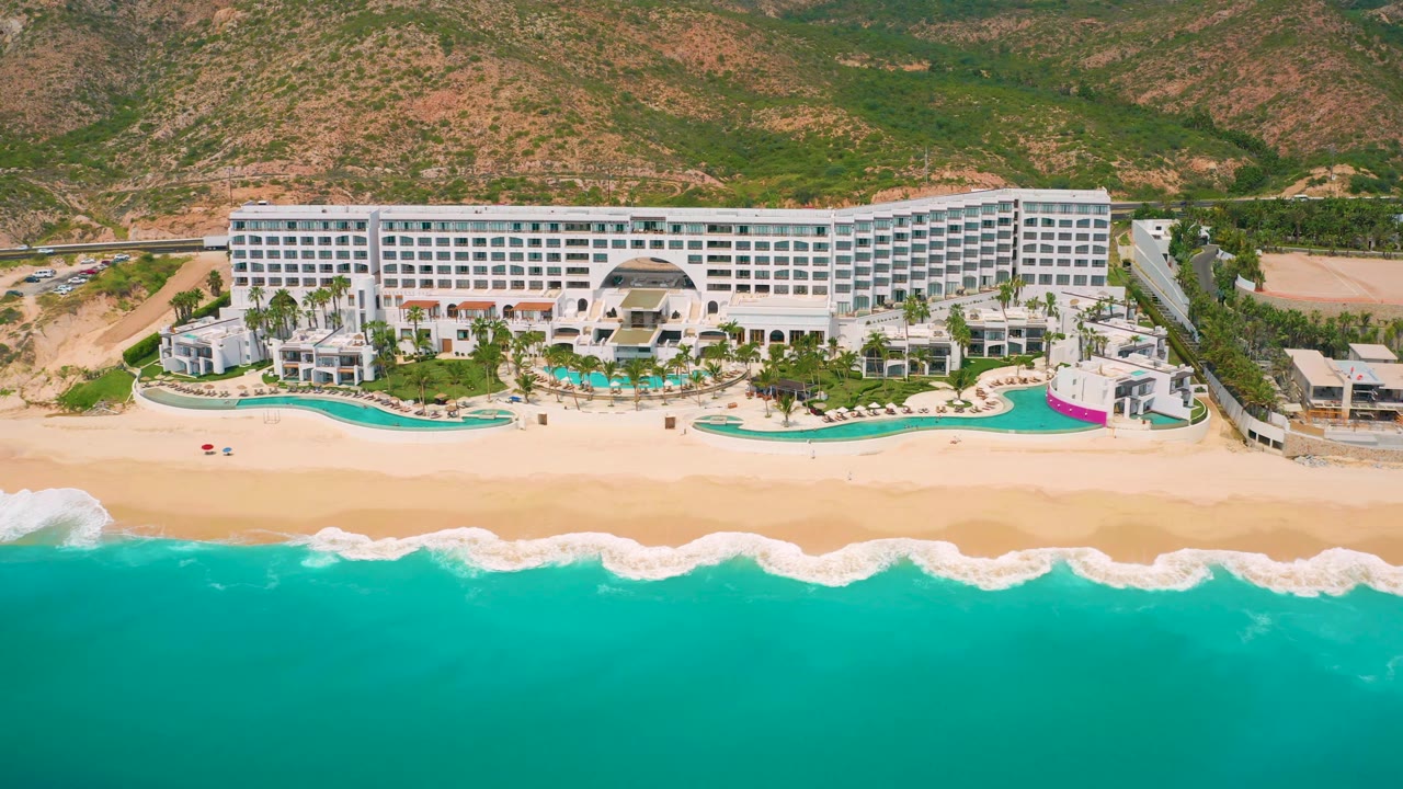 Wide aerial frame at Secrets Marquis Los Cabos showing a broad white hotel facade, bright turquoise water across the lower half, tan beach, pools, palm trees, hills, and tiny visible people on the grounds.