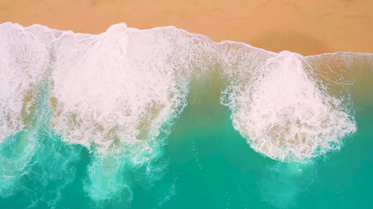 Aerial frame of broad white foam spreading over shallow blue-green water beside tan sand at Secrets Marquis Los Cabos, Mexico, with no visible people.