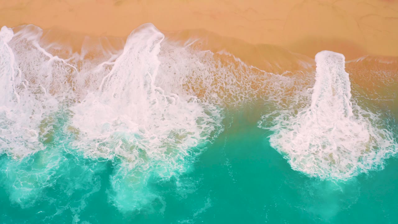 Top-down beach frame with two separated white foam plumes reaching from turquoise water onto tan sand at Secrets Marquis Los Cabos, Mexico, with no visible people.