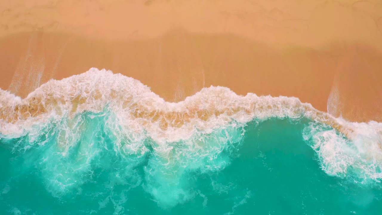 Top-down coastal frame at Secrets Marquis Los Cabos, Mexico, where a broader band of white foam spreads over aquamarine water beside golden sand, no visible people.