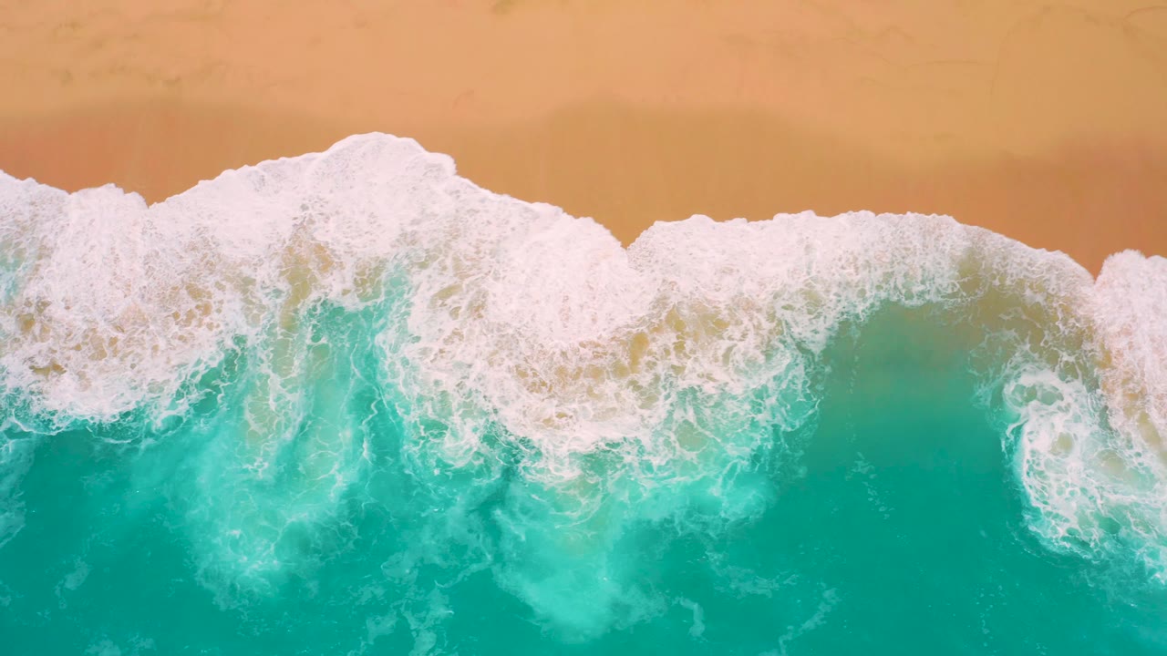 Overhead frame at Secrets Marquis Los Cabos, Mexico, showing dense white surf washing over pale green shallow water below a clean strip of golden sand, no visible people.