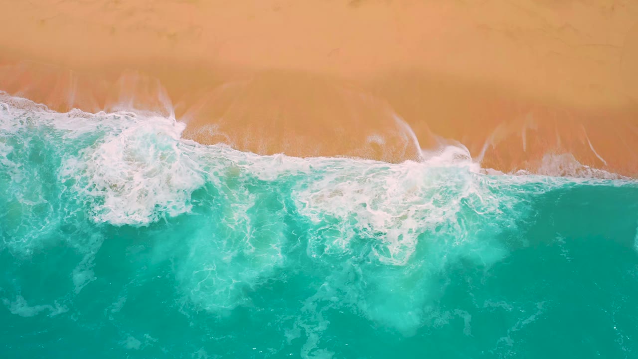 Aerial beach frame at Secrets Marquis Los Cabos, Mexico, with white foam spreading wider across turquoise water below the orange sand, with no visible people.
