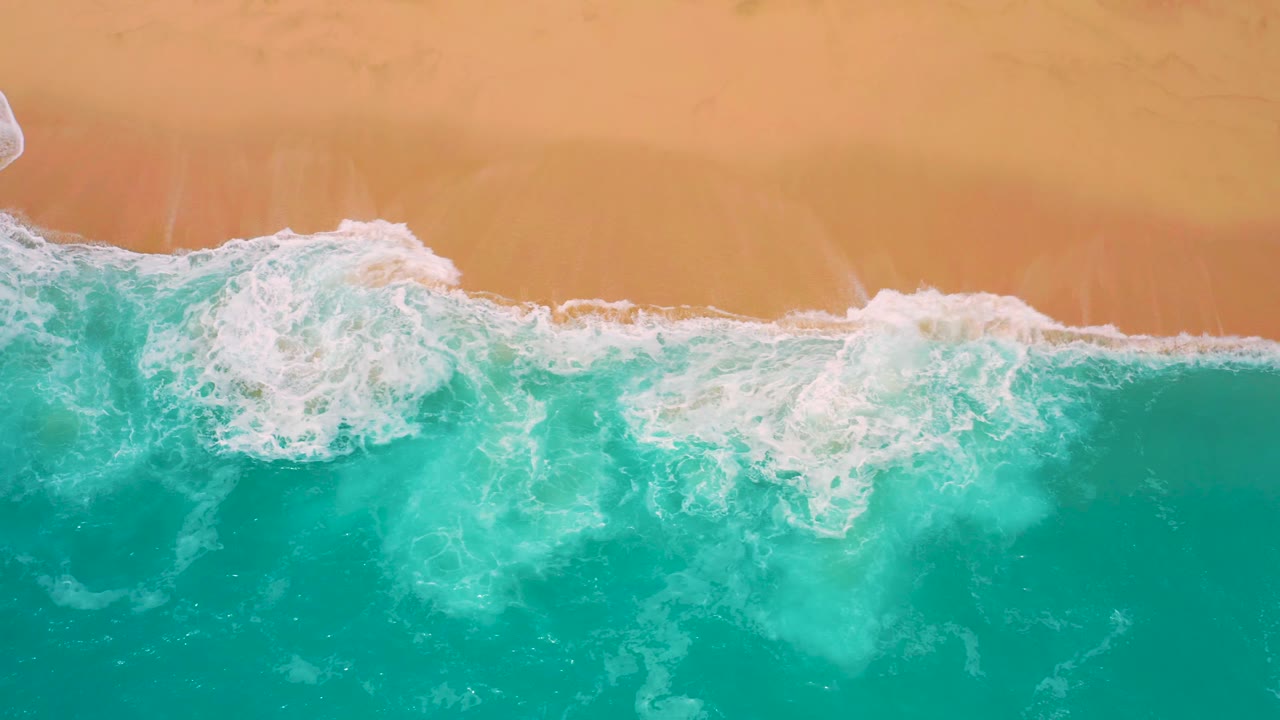 Overhead view at Secrets Marquis Los Cabos, Mexico, with a breaking wave curling along the sand and pale foam drifting into green-blue water, with no visible people.