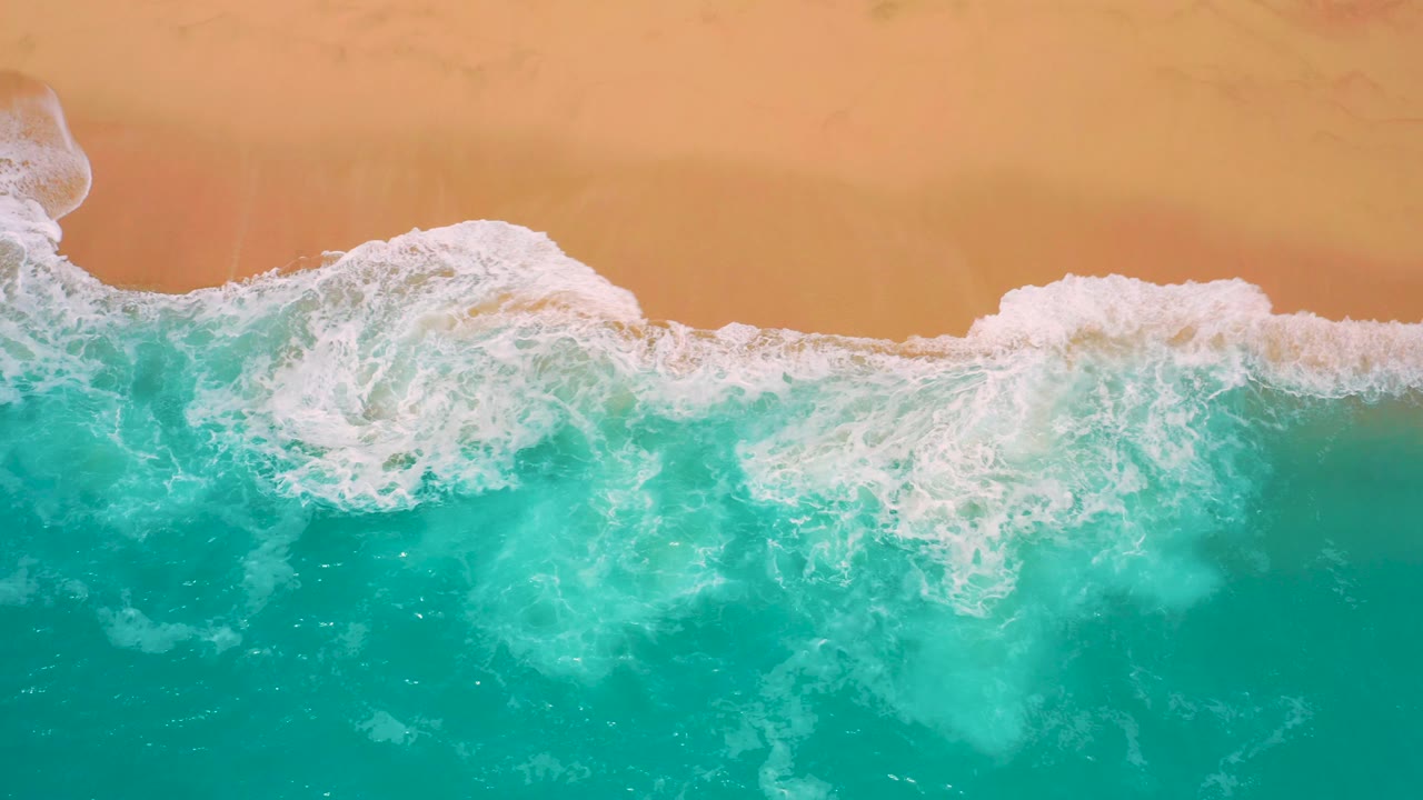 Top-down Secrets Marquis Los Cabos, Mexico beach view with a foamy wave pushing diagonally across clear blue-green water toward orange sand, with no visible people.
