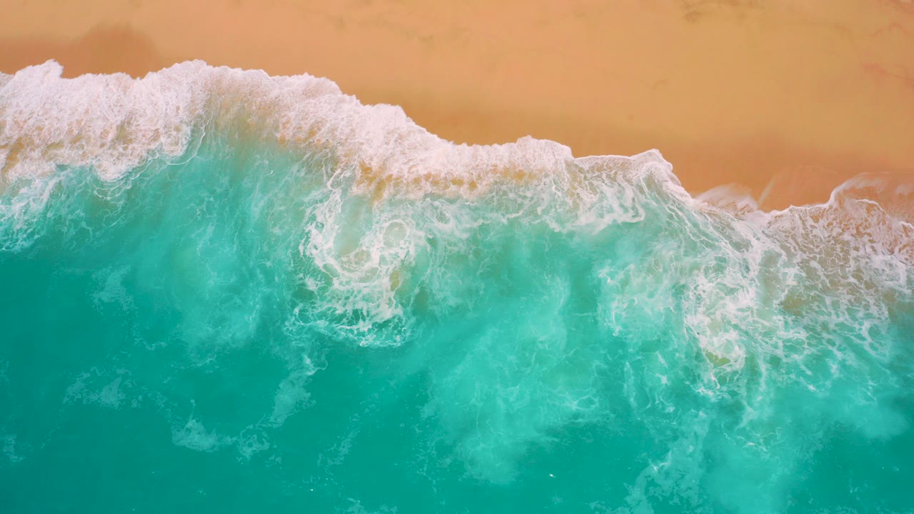 Overhead view at Secrets Marquis Los Cabos, Mexico, with teal water filling the lower frame, white foam along the upper surf line, tan sand beyond it, and no visible people.
