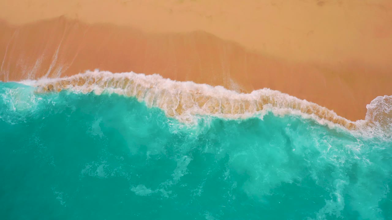 A larger white wave curls across the upper edge of turquoise water toward orange-red sand in the Mexico beach aerial, with no visible people or structures.