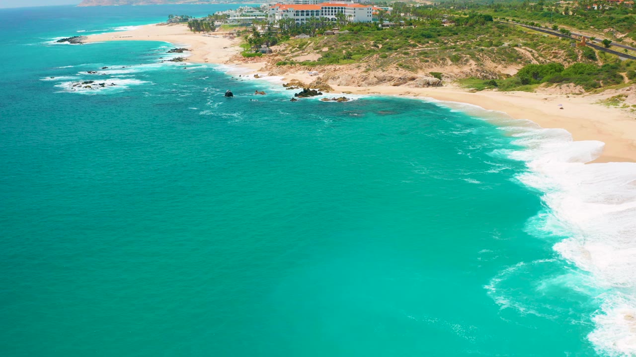 Aerial frame at 0:01 over clear blue water at Secrets Marquis Los Cabos, Mexico, with the surf line shifted along the right beach edge and no visible people.