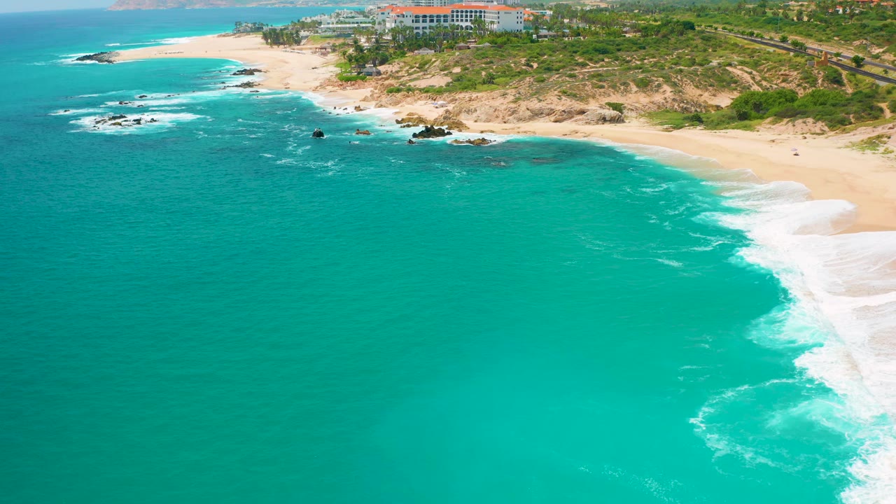 At 0:02, the aerial view shows bright turquoise ocean, a white band of surf on the right, sandy shoreline, low dunes, distant resort buildings, and no visible people at Secrets Marquis Los Cabos, Mexico.