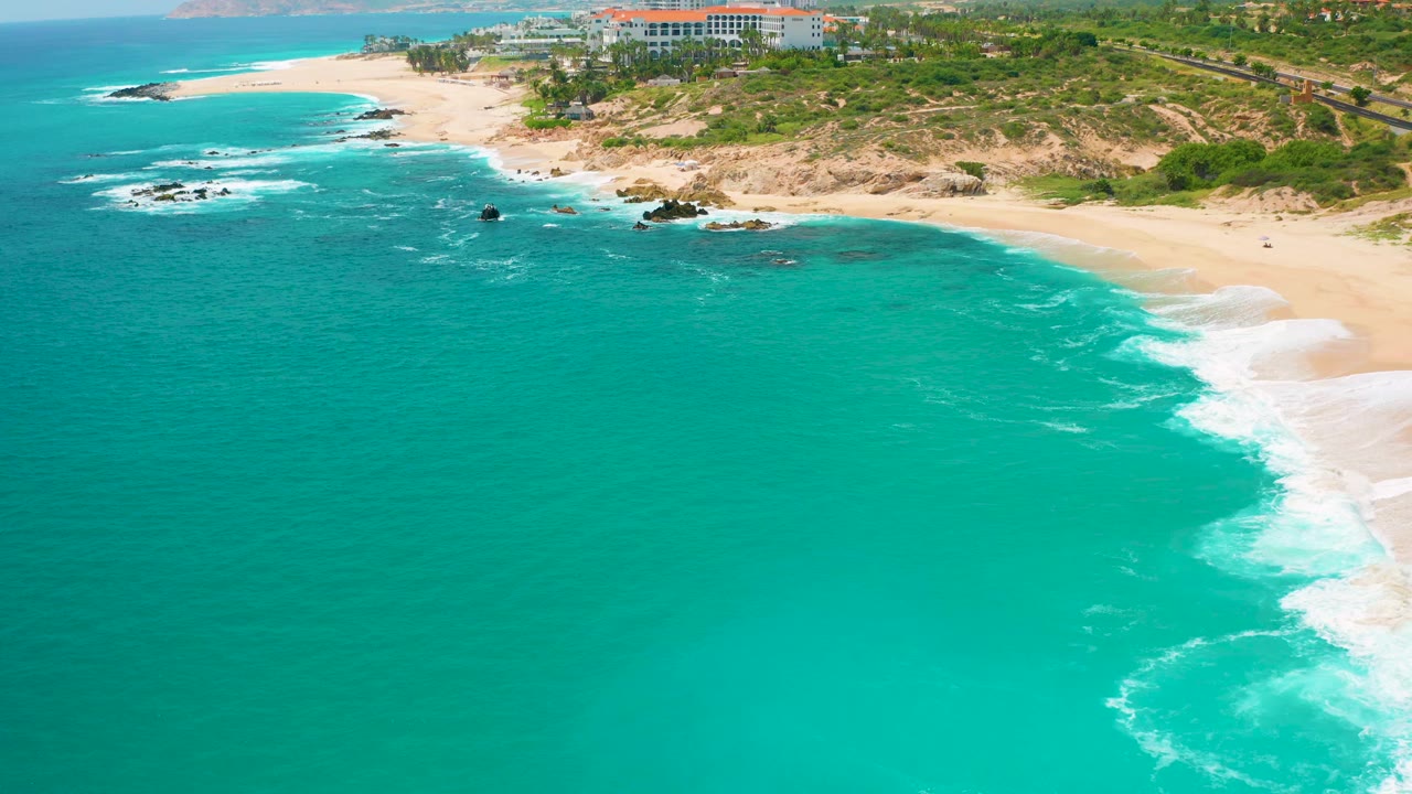 Mid-clip aerial frame over Secrets Marquis Los Cabos, Mexico, showing turquoise sea, dark rock clusters near the beach, pale sand, green coastal dunes, resort buildings, and no visible people.