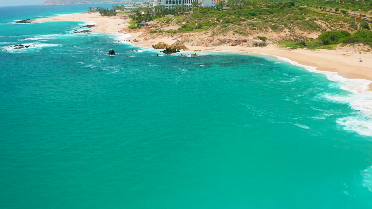 Aerial view at Secrets Marquis Los Cabos, Mexico, with open turquoise water dominating the frame, small white wave lines near shore, dark rocks offshore, and no visible people.