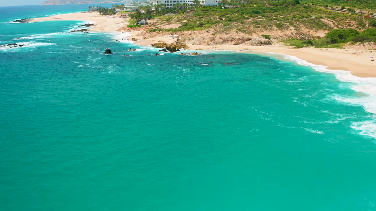 Secrets Marquis Los Cabos, Mexico coastline seen from above, with a larger sweep of turquoise water, surf brushing the sand at right, low green terrain above the beach, and no visible people.