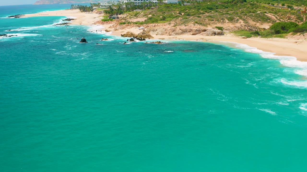 Aerial coastal frame from Secrets Marquis Los Cabos, Mexico, showing the beach curve, foamy surf at right, scattered dark rocks near shore, hillside vegetation, and no visible people.