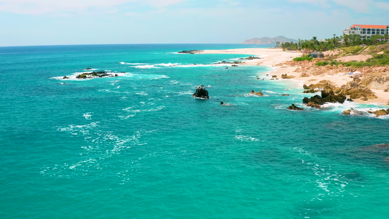 Wide elevated view of turquoise ocean, dark rocks, sandy beach, palms, and a distant red-roofed resort at Secrets Marquis Los Cabos, Mexico, with no visible people.