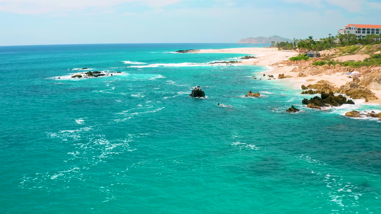 Wide 4K frame looking across open blue-green water toward a sandy, rocky coast in Mexico, with small surf bands, scattered dark rocks, and no visible people.