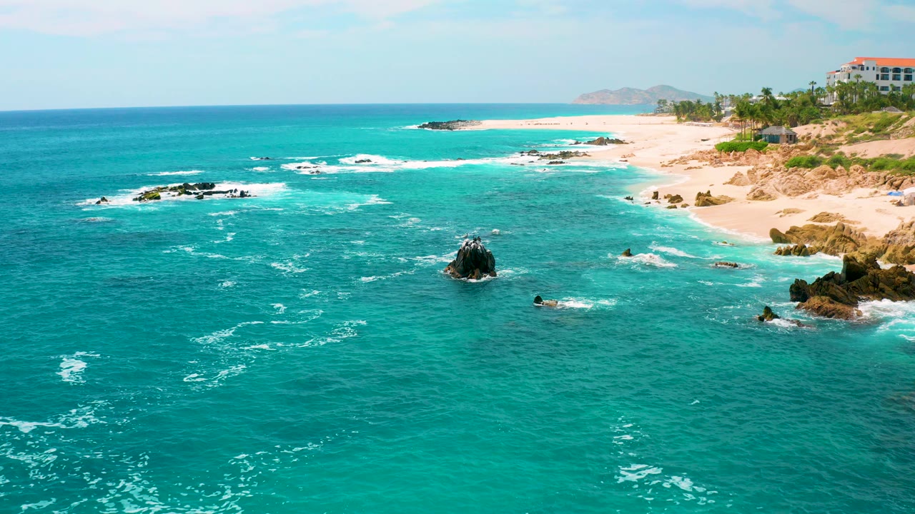 Aerial coastal frame at Secrets Marquis Los Cabos, Mexico, with turquoise water in the foreground, dark rocks breaking the surf, pale sand to the right, resort buildings at the upper right, and no visible people.