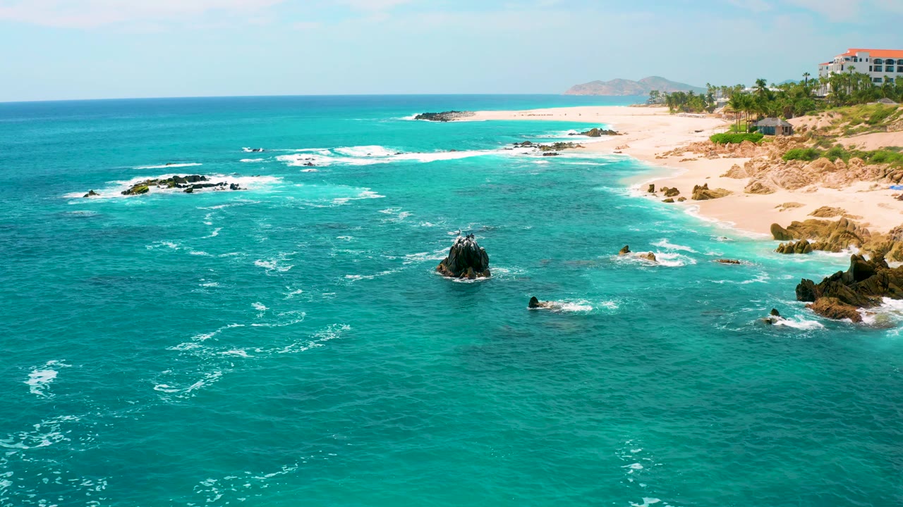 Wide aerial view at Secrets Marquis Los Cabos, Mexico, with the coastline running along the right side, teal ocean covering most of the frame, surf lines near black rocks, and no visible people.