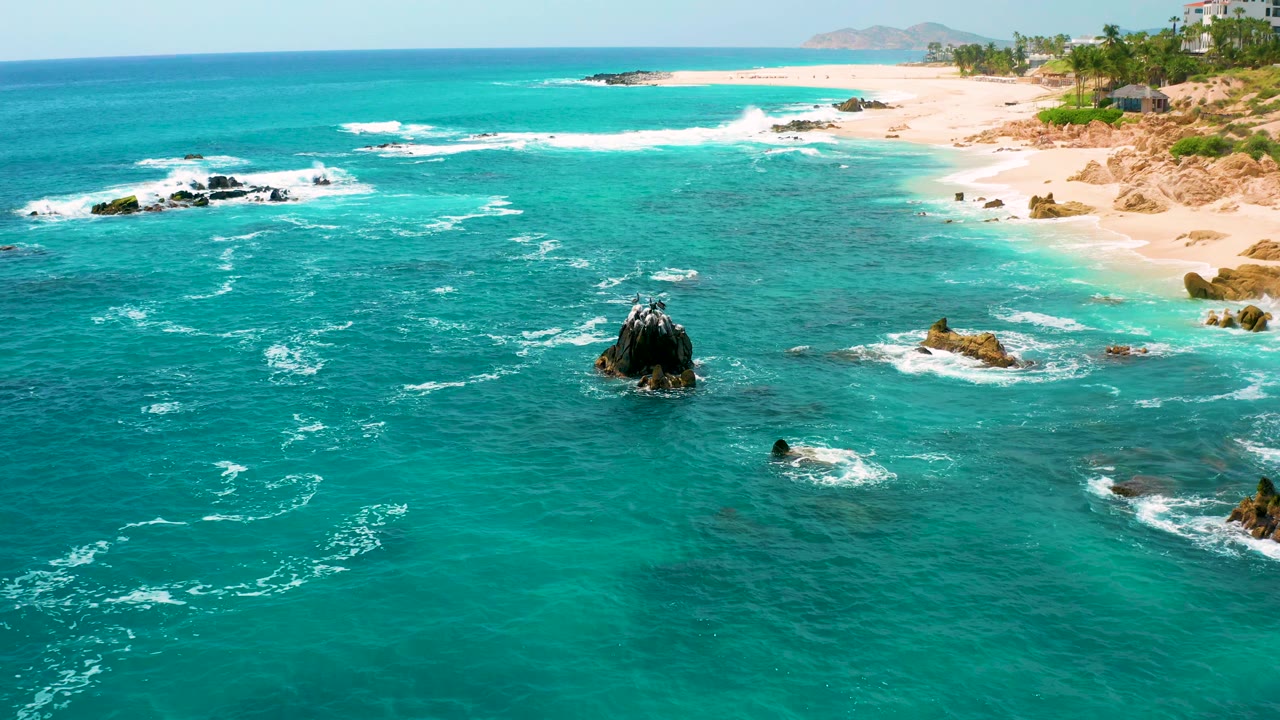 Drone aerial over Secrets Marquis Los Cabos, Mexico showing bright blue-green water, a central dark rock, smaller surf-washed rocks, pale sand, and no visible people.