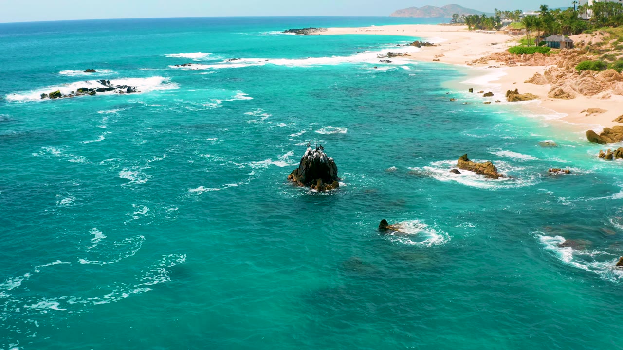 Wide aerial frame at Secrets Marquis Los Cabos, Mexico with turquoise sea in the foreground, foamy wave trails around rocks, sandy beach along the upper right, and no visible people.