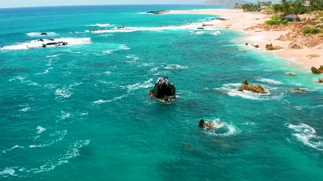 Aerial shoreline view with surf bands, dark rocks, and beach.