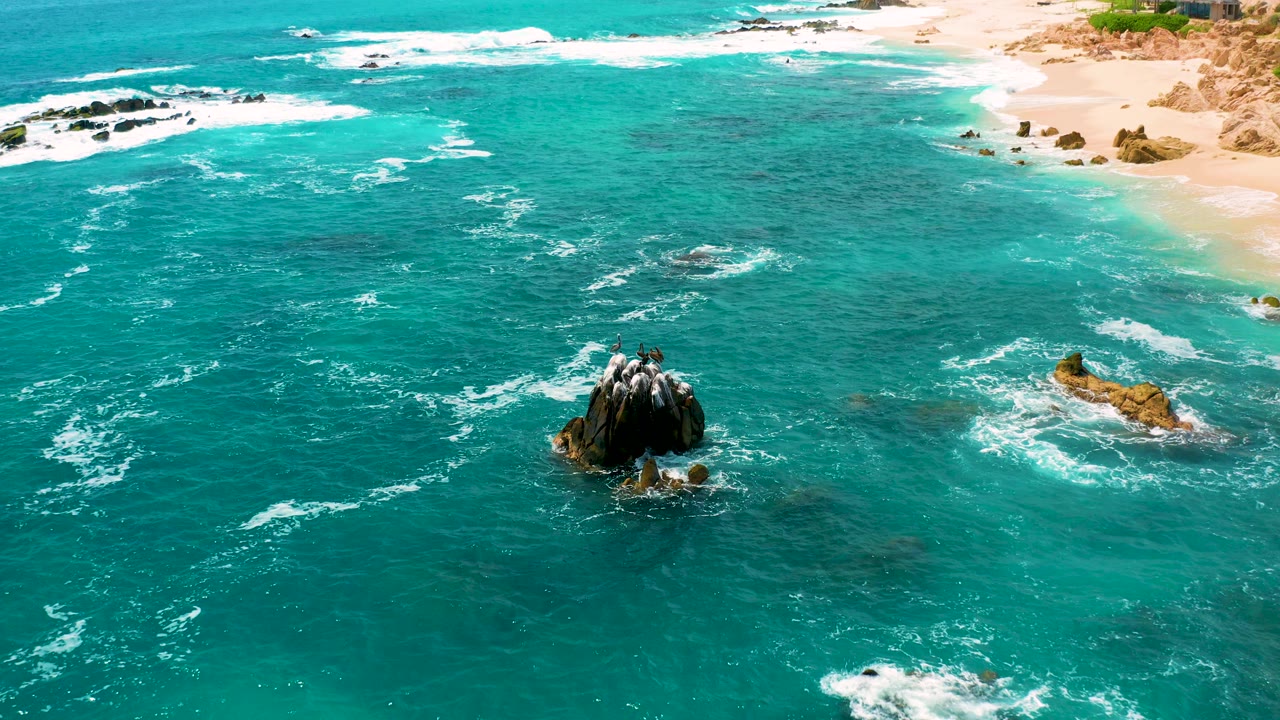 Aerial view at Secrets Marquis Los Cabos, Mexico, with turquoise water surrounding a dark rock outcrop, sandy beach along the upper edge, and no visible people.