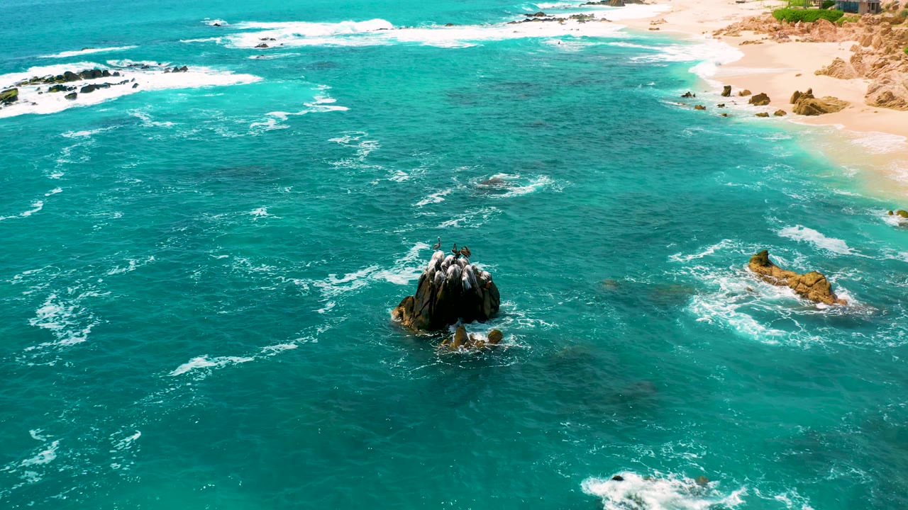 Blue-green ocean surrounds a protruding rock cluster with white surf lines and beach in the background.