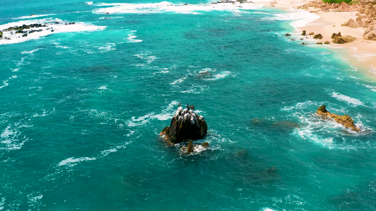 At Secrets Marquis Los Cabos, Mexico, the aerial view shows the rock outcrop slightly lower in frame, white wave wash spreading across turquoise water, sand at the top, and no visible people.