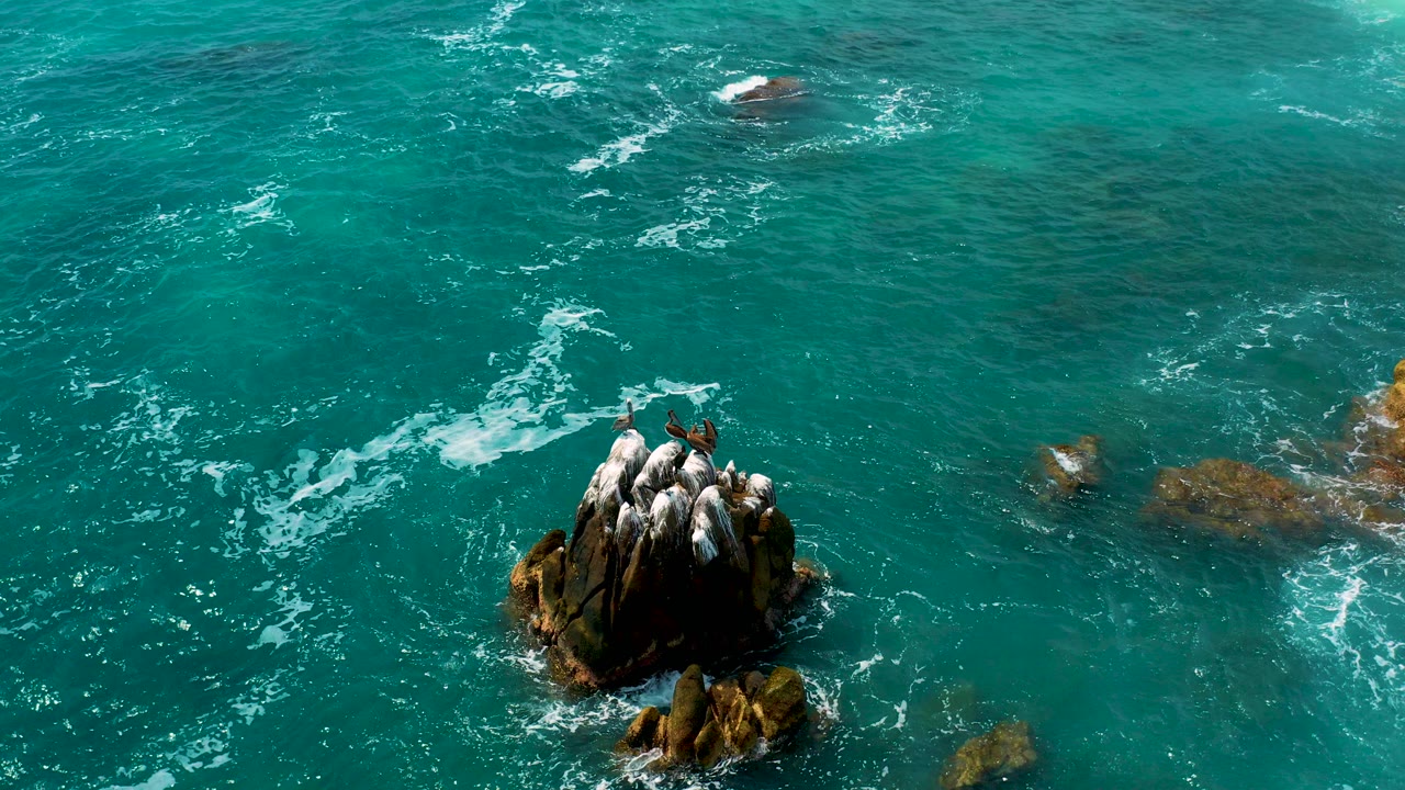 Overhead view of turquoise water swirling around a rocky outcrop in Los Cabos, Mexico, with white foam trails and no visible people.