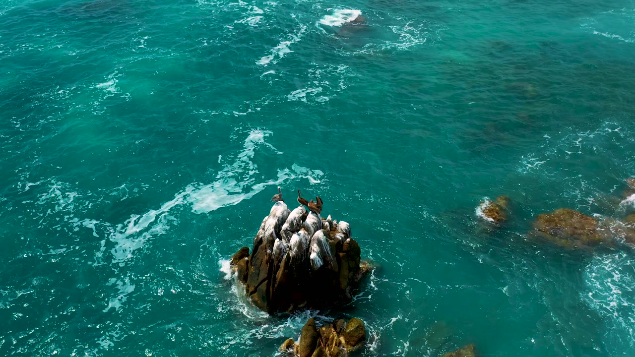 High overhead view in Los Cabos, Mexico, where the rock cluster sits right of center amid turquoise water, thin foam lines, and no visible people.