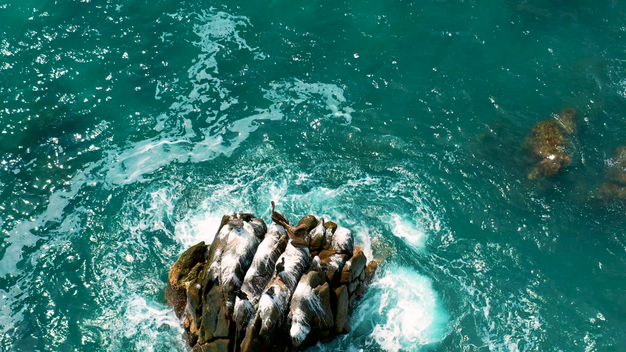 Overhead view at Secrets Marquis Los Cabos, Mexico, with turquoise water and white foam surrounding a dark rock formation near the lower frame, no visible people.